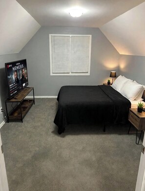 A bedroom with a bed, a TV on a stand, a window with blinds, and a nightstand with a lamp and a plant. - 3-bedroom house  2 cozy rooms in peaceful Harvey with shared living space (Harvey)