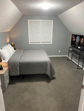A bedroom with a bed, a nightstand, a lamp, a TV, and a window with blinds. - Cozy 2-bedroom house in peaceful Harvey with AC (Harvey)