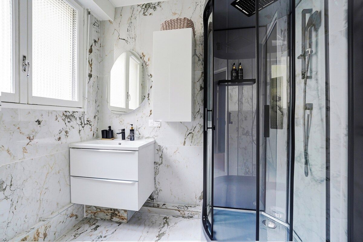 A modern bathroom with a marble wall, a white vanity, a glass shower enclosure, and a large mirror.