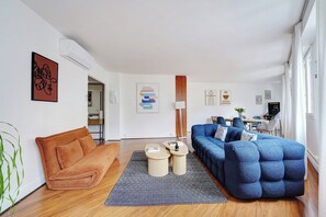 A modern living room with a sofa, a coffee table, and a blue sectional. There is a dining area in the background with a table and chairs. - Contemporary Gem – Eiffel Tower & Metro Nearby (Paris)