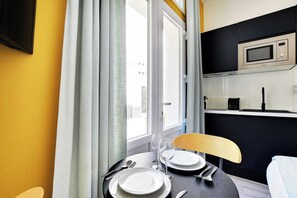 Ein Essbereich mit Tisch für zwei Personen, einem Stuhl, einem Fenster mit Vorhängen und einer Küchenzeile inklusive Mikrowelle.