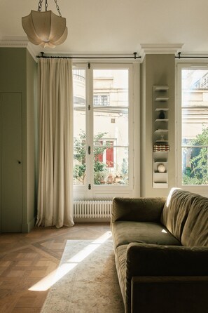 A room with a sofa, a radiator, a window with curtains, and a hanging light fixture. - Curated Home for an Unforgettable Experience (Paris)