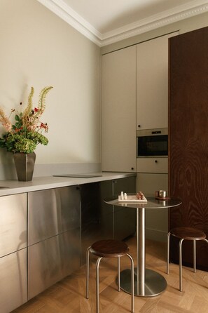 A modern kitchen with stainless steel cabinets, a round dining table, two stools, and a potted plant. - Curated Home for an Unforgettable Experience (Paris)
