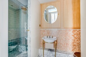 A bathroom with a glass shower enclosure, a marble-tiled floor, a sink with gold fixtures, and a wooden vanity cabinet. - Parisian Escape in Central Le Marais (Paris)
