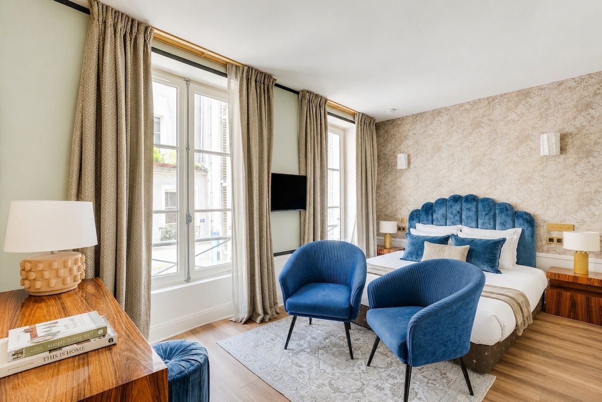 Un dormitorio con una cama, dos sillones azules, una mesita de madera con una lámpara y una ventana con cortinas.