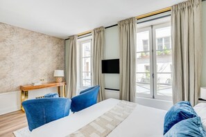 Et hotelværelse med en seng, to blå stole, et fjernsyn og et vindue med gardiner.