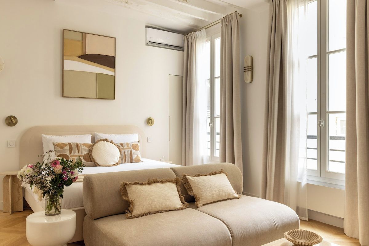 Ein Schlafzimmer mit einem Bett, einem Sofa, einem Nachttisch mit Blumen und einem Fenster mit Vorhängen.
