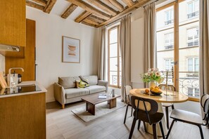 A modern living room with a sofa, coffee table, dining table, and chairs. There is a window with curtains, a kitchen area with a sink and cabinets, and a framed picture on the wall. - Chic 1BR only Steps away from the Louvre (Paris)