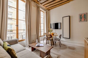 A furnished living room with a sofa, a wooden coffee table, a dining table with chairs, a flat-screen TV mounted on the wall, and a balcony with a railing. - Chic 1BR only Steps away from the Louvre (Paris)