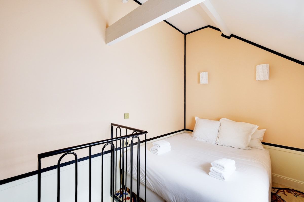 A hotel room with a bed, white linens, a black metal railing, and a small window.