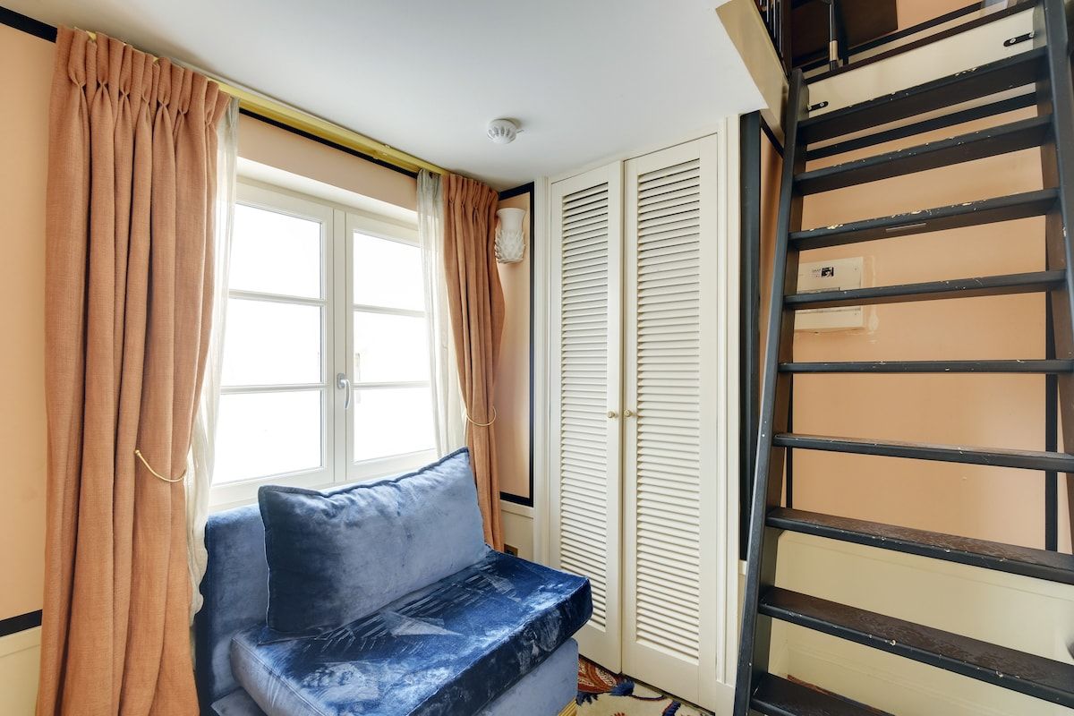 Een kamer met een bank, een raam met gordijnen, een deur met verstelbare luiken en een metalen ladder, zwart van kleur.