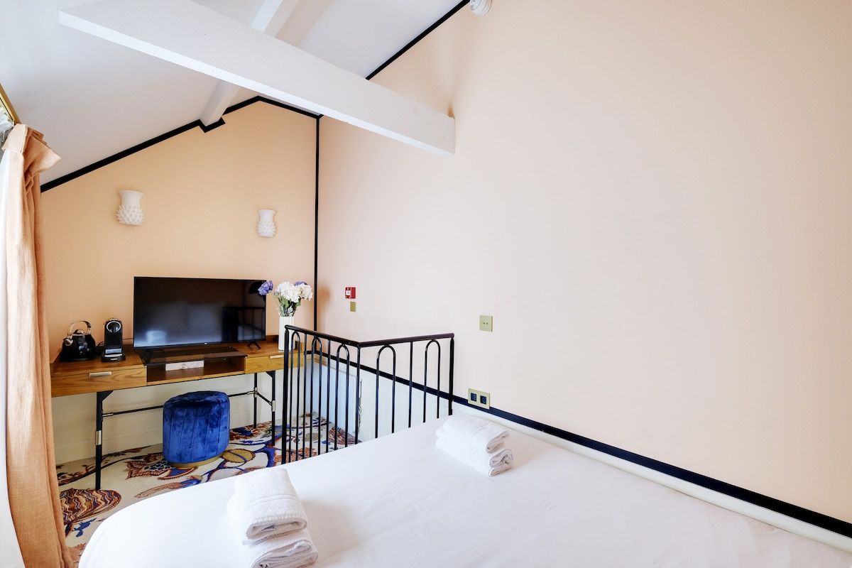 Une chambre d’hôtel avec un lit, un bureau avec une télévision, un petit tabouret et un pouf bleu.