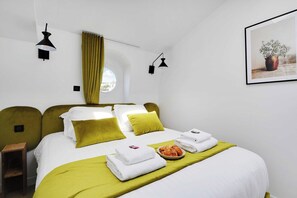 A hotel room with a bed, two bedside lamps, a window with curtains, a framed picture on the wall, and a small table with towels and a plate of pastries. - Cosy Cocoon Nestled in Vibrant 11th - Voltaire (Paris)