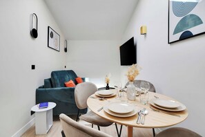 A modern dining area with a round table set for four, a blue sofa, and a flat-screen TV mounted on the wall. - Cosy Cocoon Nestled in Vibrant 11th - Voltaire (Paris)