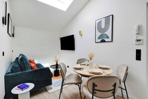 A modern living room with a sofa, a dining table set for four, and a flat-screen TV mounted on the wall. - Cosy Cocoon Nestled in Vibrant 11th - Voltaire (Paris)
