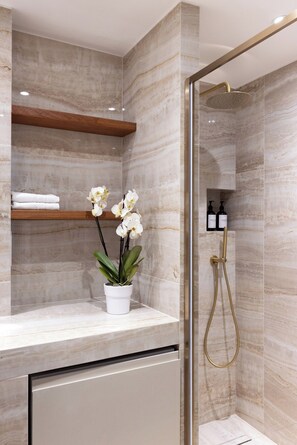 A modern bathroom with marble walls, a glass shower enclosure, a white sink with a potted orchid, and neatly folded towels on a shelf. - Brand New Flat in Central Marais - Walk Everywhere (Paris)