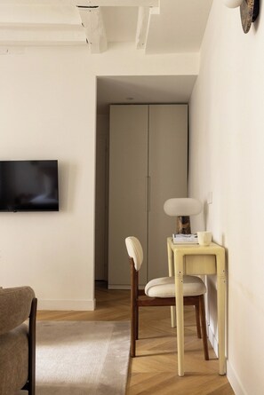 A minimalist living room with a flat-screen TV mounted on the wall, a wooden chair, a small table with a lamp, and a door leading to another room. - Brand New Flat in Central Marais - Walk Everywhere (Paris)