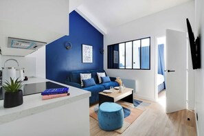 A modern living space with a blue accent wall, a sofa, a coffee table, and a kitchen area with a sink and countertop. - Sleep Above the Parisian Clouds - 1BR - Voltaire (Paris)