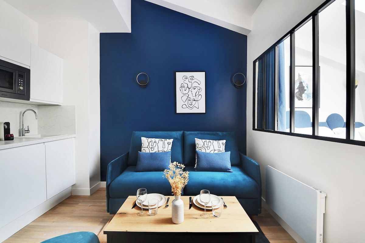 Un salon moderne avec un mur d’accent bleu, un canapé agrémenté de coussins à motifs, une table basse avec un vase, et un coin cuisine avec des meubles blancs et un four à micro-ondes.