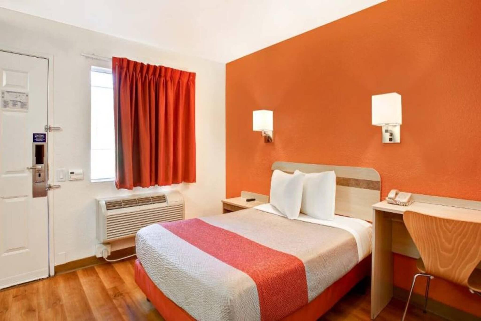 A hotel room with a bed, bedside tables, a telephone, an air conditioning unit, and a window with curtains.