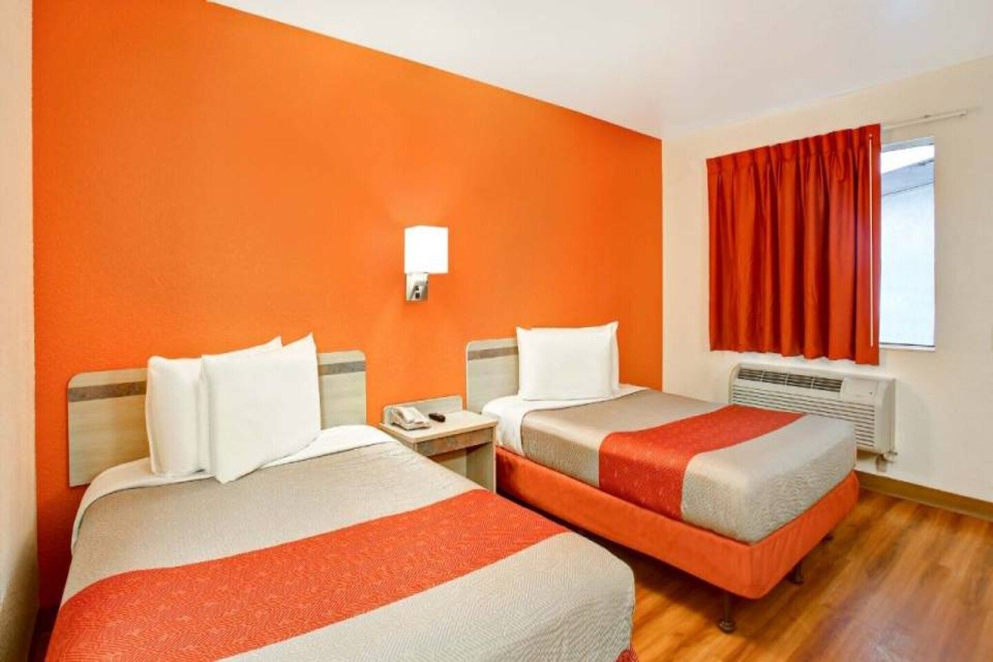 Ein Hotelzimmer mit zwei Betten, einem Nachttisch, einem Fenster mit roten Vorhängen und einer Wandleuchte.
