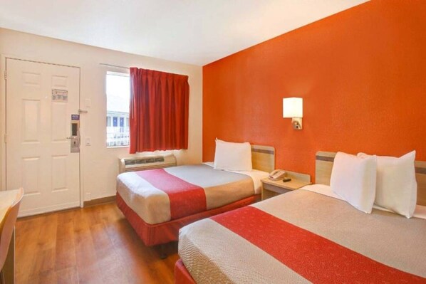 A hotel room with two beds, a window with red curtains, a wall-mounted light, and a door with a lock.
