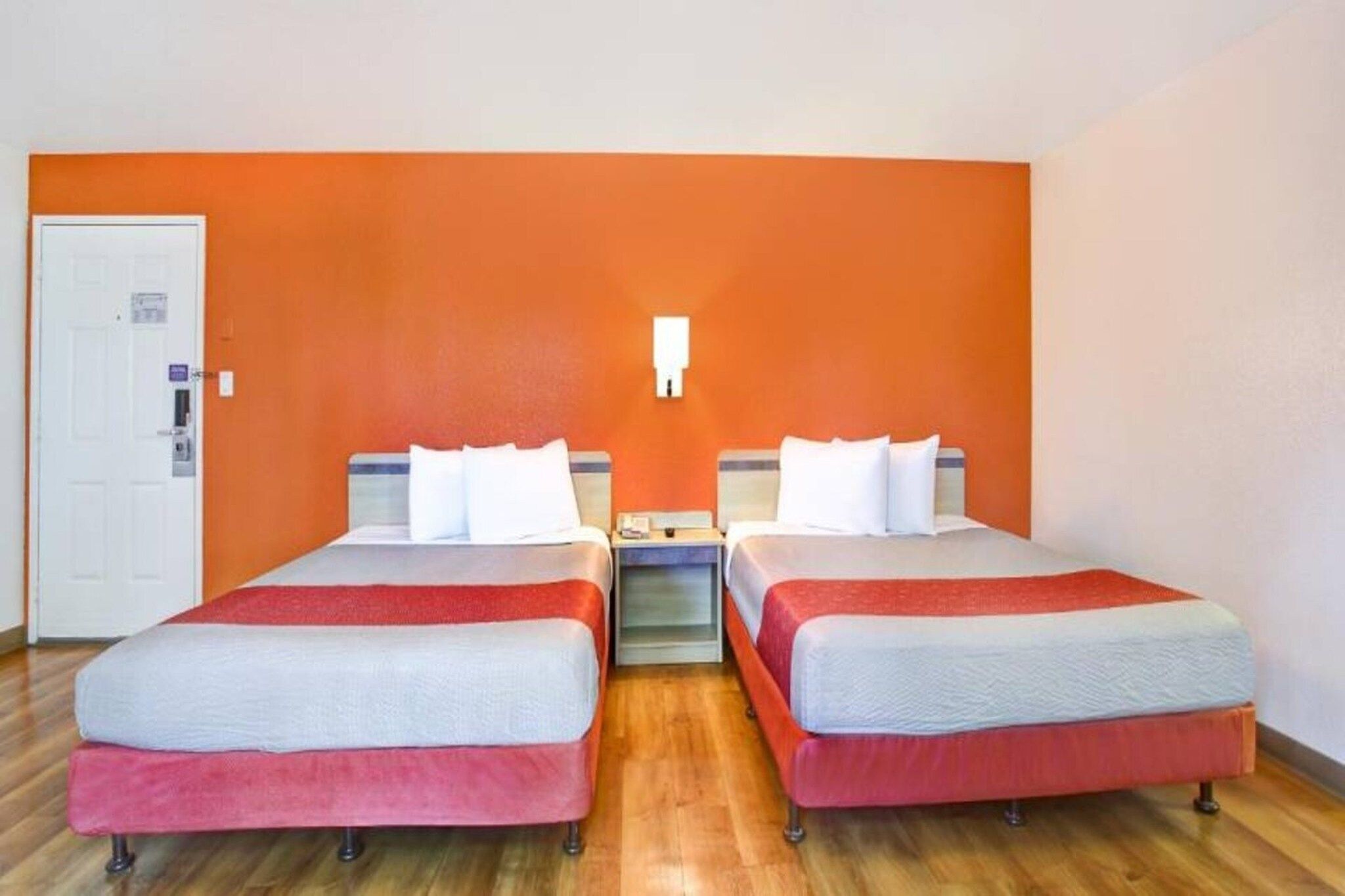 A hotel room with two beds, a nightstand, a white door, and an orange wall.