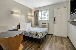 Ein kleiner Raum mit einem Bett, einem Schreibtisch, einem Stuhl, einem Fenster mit Vorhängen und einer Tür.