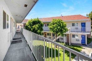 Een gang met een balkon dat uitkijkt over een rij tweelaags gebouwen met rode dakpannen en blauwe deuren.