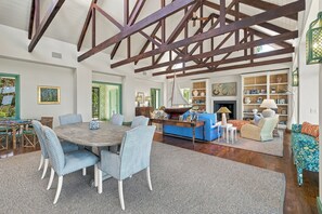 A spacious living area with a high ceiling, exposed wooden beams, a round dining table with chairs, a blue sofa, and a fireplace.