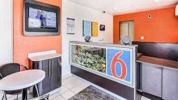 Zona de recepción de hotel con mostrador, televisor montado en la pared y un área de estar con mesa y sillas.