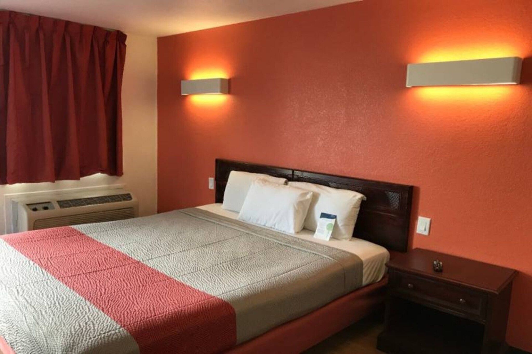A hotel room with a bed, a nightstand, a wall-mounted heater, and a window with curtains.