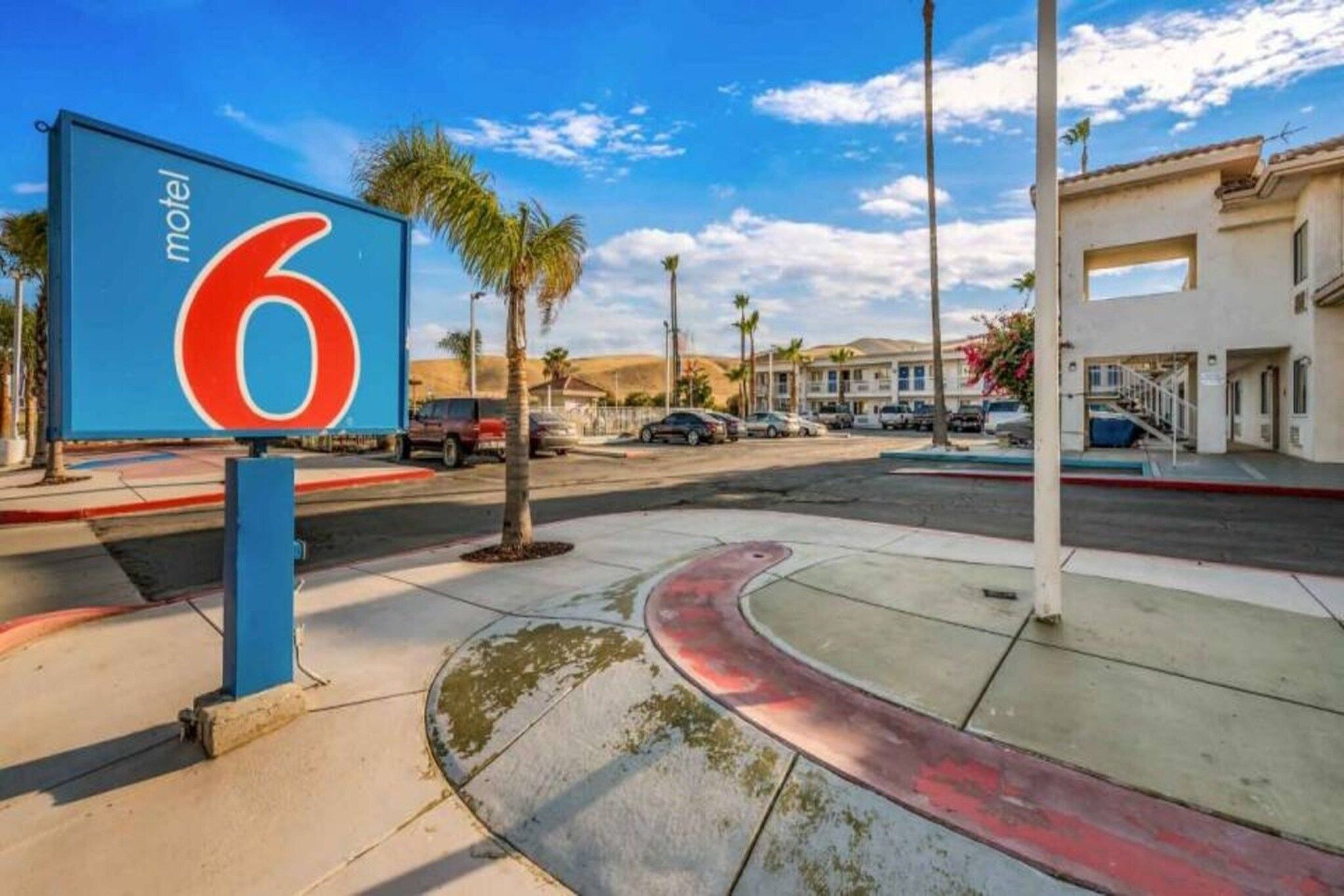 A motel sign with the number 6, a parking lot with several cars, palm trees, and a building with a flat roof in the background.