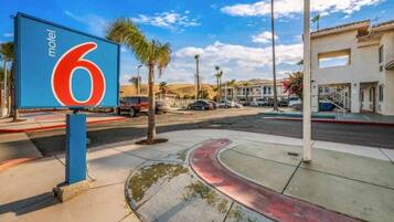 A motel sign with the number 6, a parking lot with several cars, palm trees, and a building with a flat roof in the background.