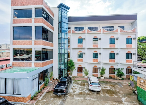 A multi-story building with a mix of brick and glass facades, a parking area with two vehicles, and a small garden area with potted plants. - Hotel Alexander (Puerto Princesa)