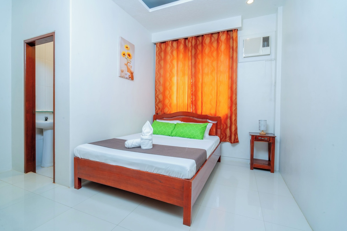A hotel room with a bed, a small bedside table, a wall-mounted air conditioning unit, and a bathroom with a sink.