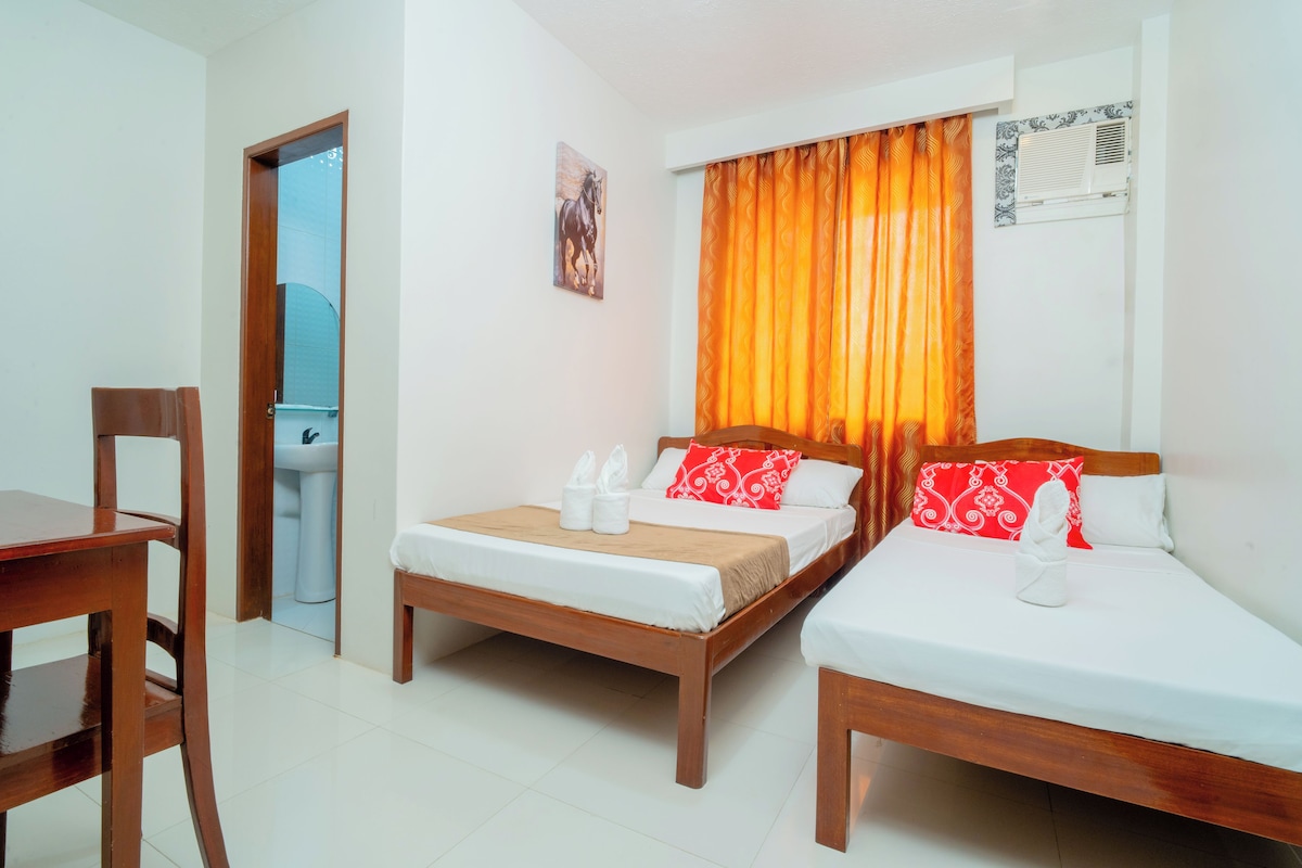 A hotel room with two single beds, a wooden desk, a chair, a bathroom with a sink, and a window with orange curtains.