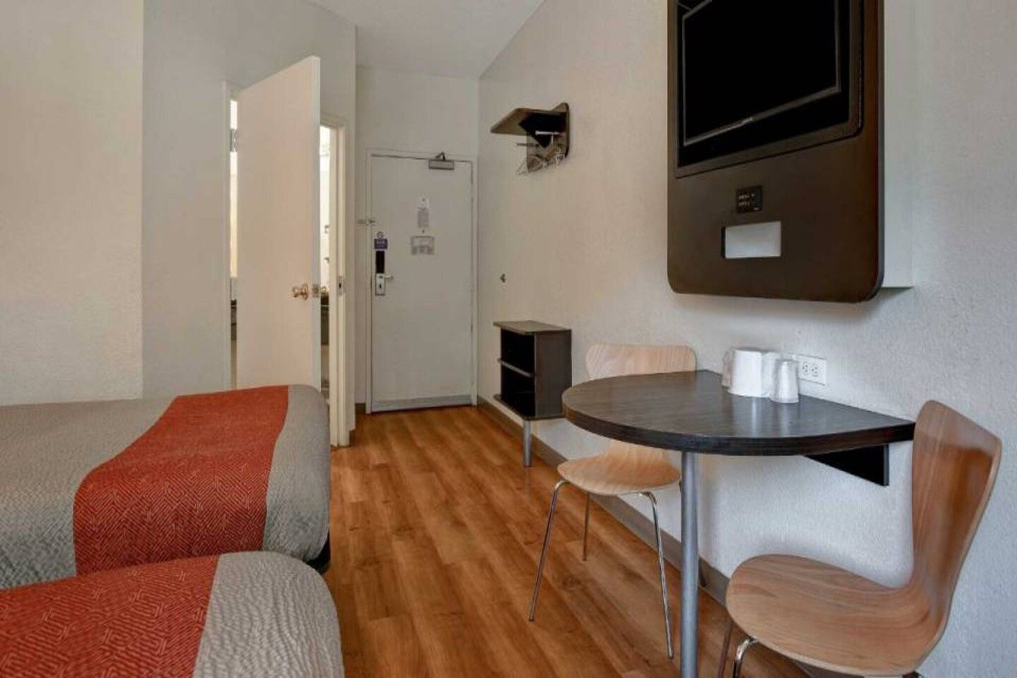 A room with a bed, a table with chairs, a television mounted on the wall, and a door leading to another room.