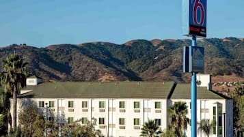 Un motel con un letrero que indica el número 6. Tiene un edificio de techos planos con múltiples ventanas, rodeado de palmeras y colinas al fondo.
