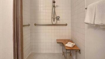 Un vestuario de ducha pequeño con azulejos, un cabezal de ducha de mano, una barra de apoyo y un banco con una toalla.