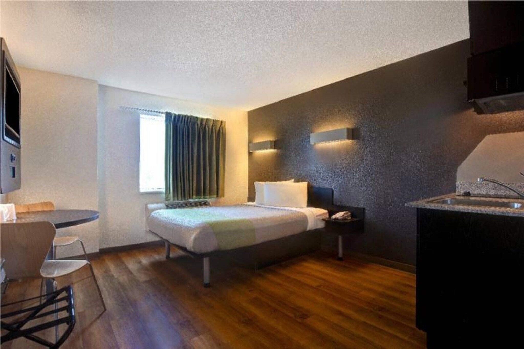 A compact hotel room with a bed, bedside table, and a small kitchenette.