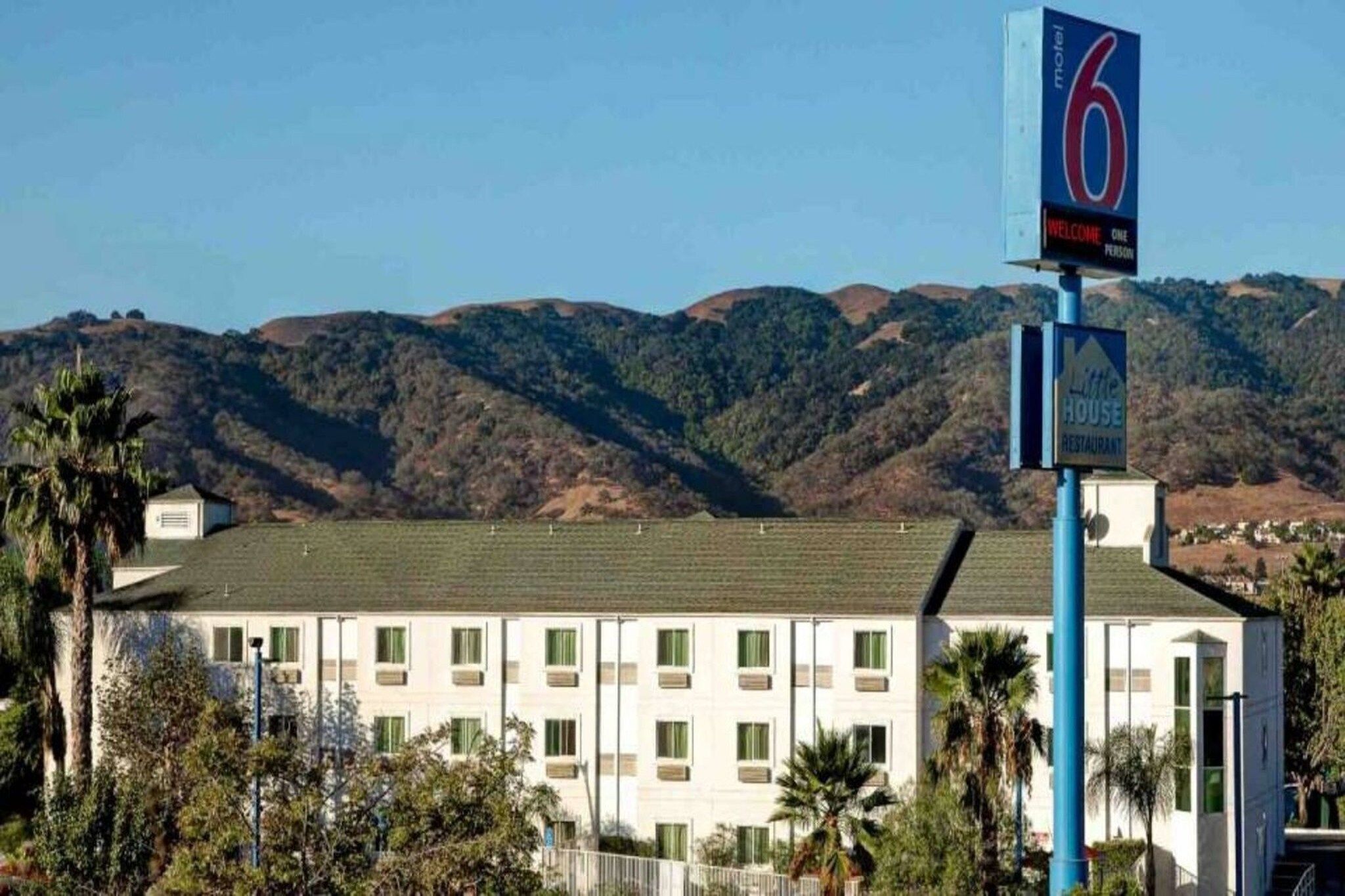A motel with a sign indicating it is number 6. It has a flat-roofed building with multiple windows and is surrounded by palm trees and hills in the background.