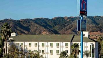 A motel with a sign indicating it is number 6. It has a flat-roofed building with multiple windows and is surrounded by palm trees and hills in the background.