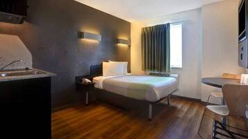 A compact hotel room with a bed, bedside table, a small desk with a chair, a window with curtains, and a kitchenette area.