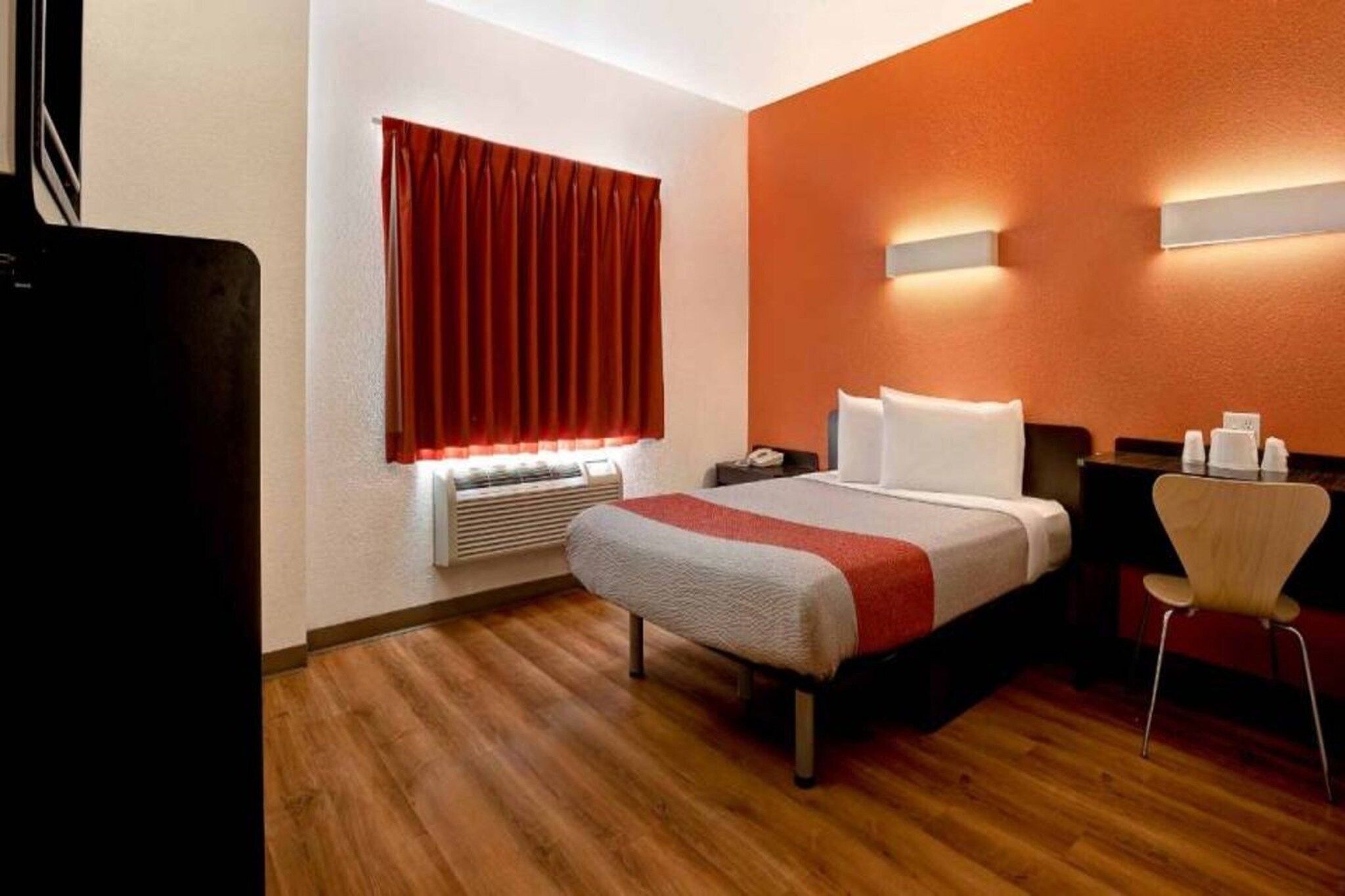 Habitación de hotel con cama, escritorio, silla, equipo de aire acondicionado y ventana con cortinas.