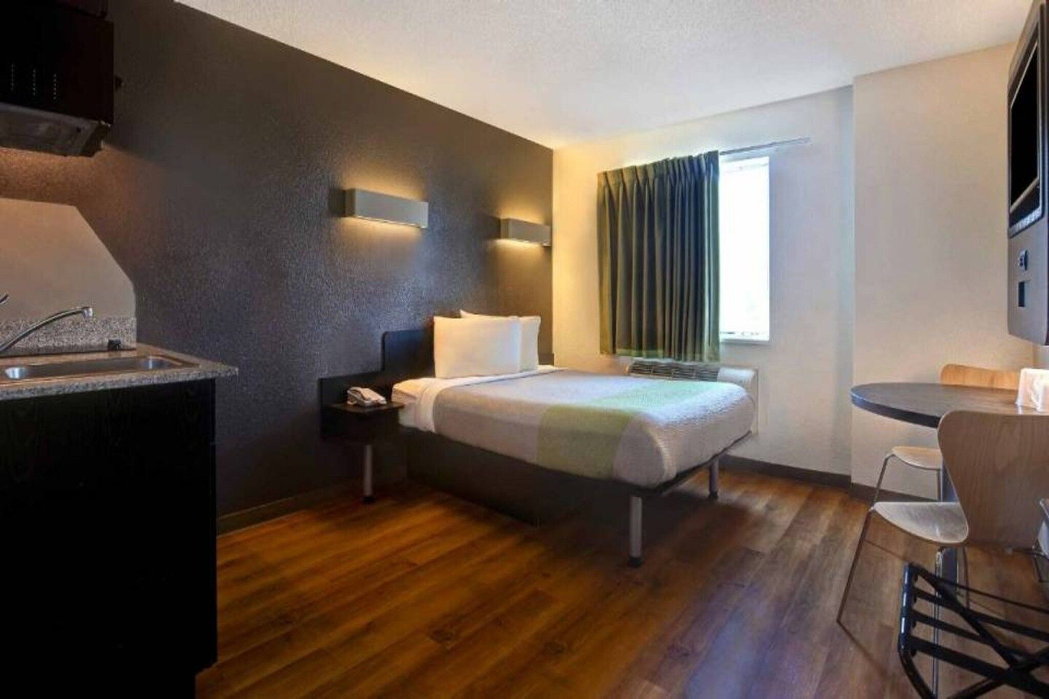 A compact hotel room with a bed, bedside table, a small desk with a chair, a window with curtains, and a kitchenette area.