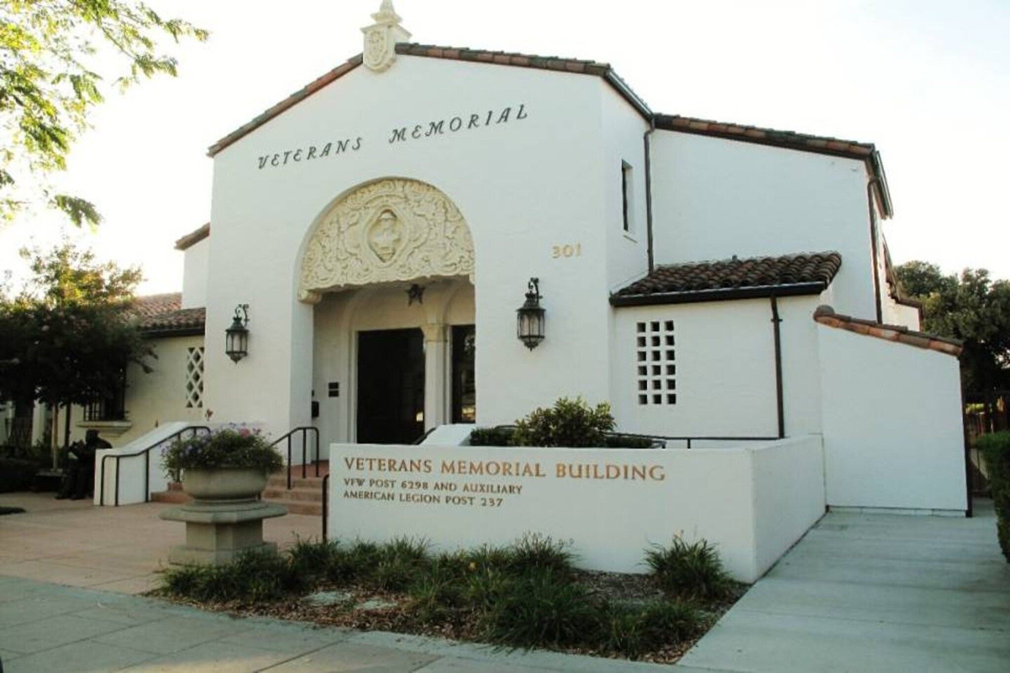 A Veterans Memorial Building with a white facade, a central arched entrance, and a small garden area with a bench.