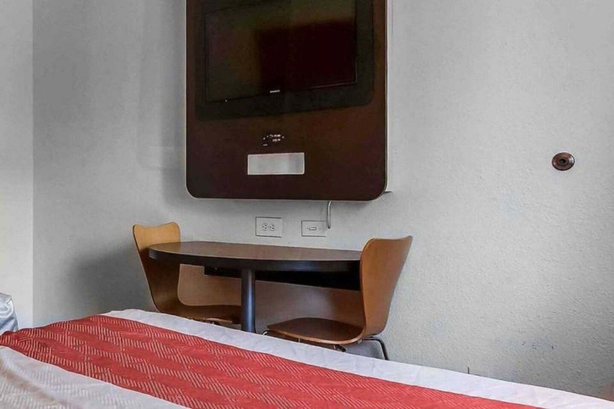 Habitación de hotel con cama, mesa, dos sillas, televisor montado en la pared y toma de corriente.