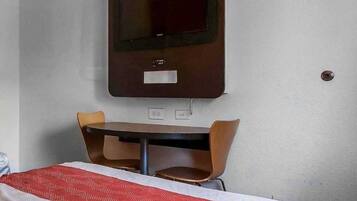 Habitación de hotel con cama, mesa, dos sillas, televisor montado en la pared y toma de corriente.
