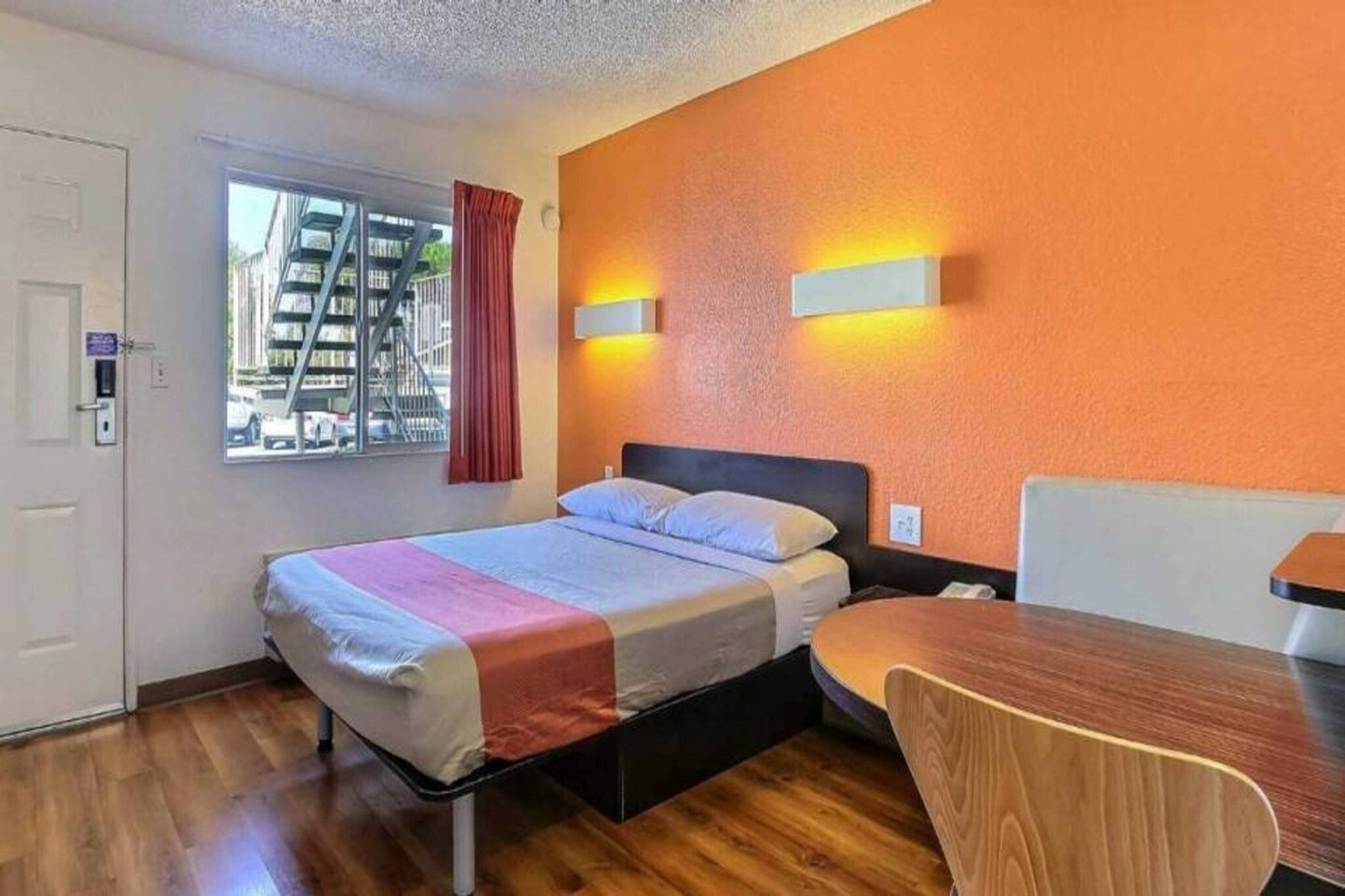 Quarto de hotel com cama, escrivaninha e cadeira. Janela com vista para edifícios.